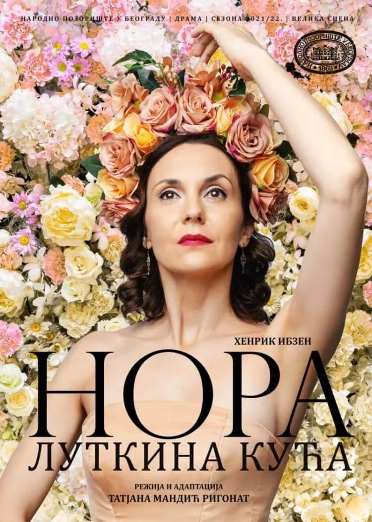 Plakat predstave Nora - Lutkina kuća: žena sa cvetnim vencem i podignutom rukom ispred zida od cveća